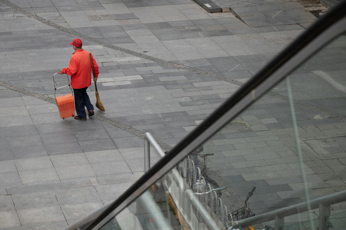 A street cleaner in a red jacket sweeping the urban pavement with a broom and cart.