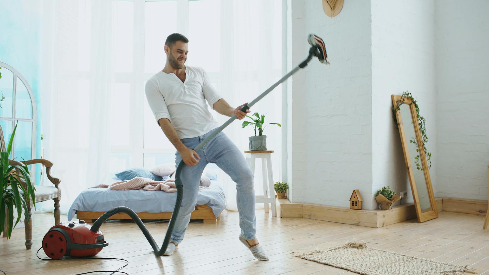 Young man energetically pretends to play guitar with a vacuum cleaner in bright bedroom.